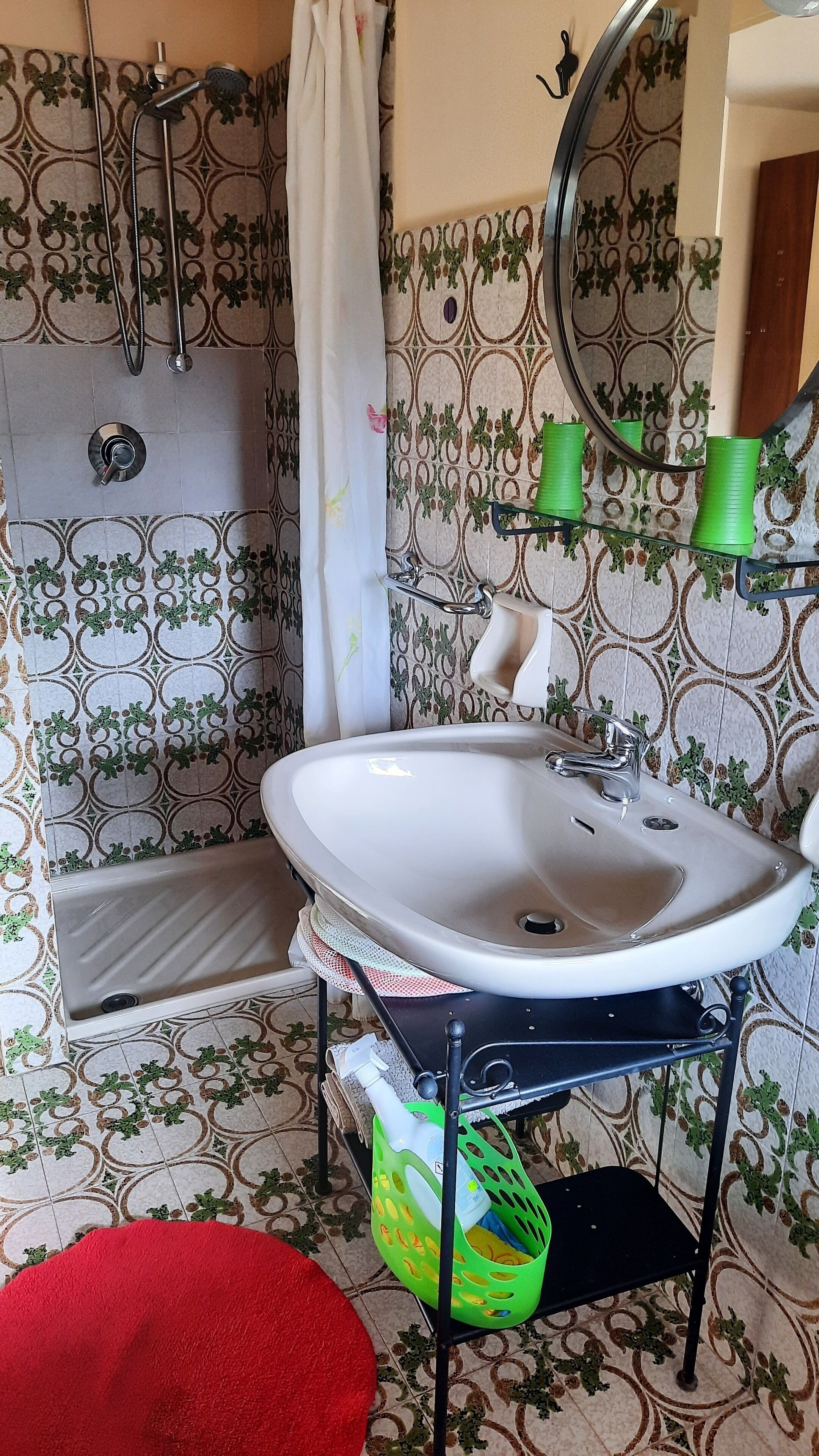 A bathroom with a white sink, shower, and toilet. There is a green basket under the sink and a red rug on the floor.
