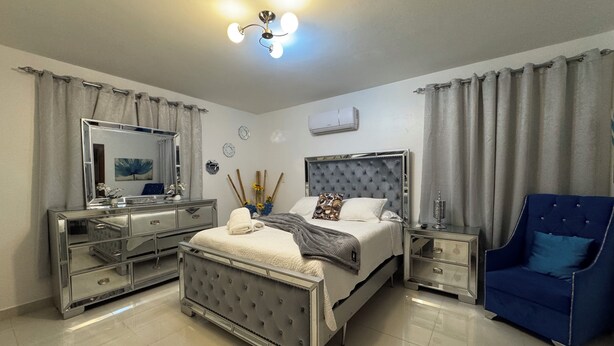 Un dormitorio moderno con una cama grande, un tocador con espejo, un sillón azul y una lámpara en el techo.