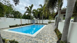 Una piscina rectangular con plataforma de baldosas de piedra, rodeada por un muro blanco y palmeras.