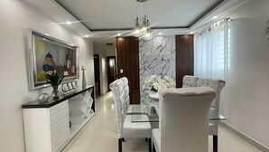 Un comedor moderno con una mesa de vidrio, sillas blancas con tapicería, una pared decorativa de mármol y una lámpara colgante.