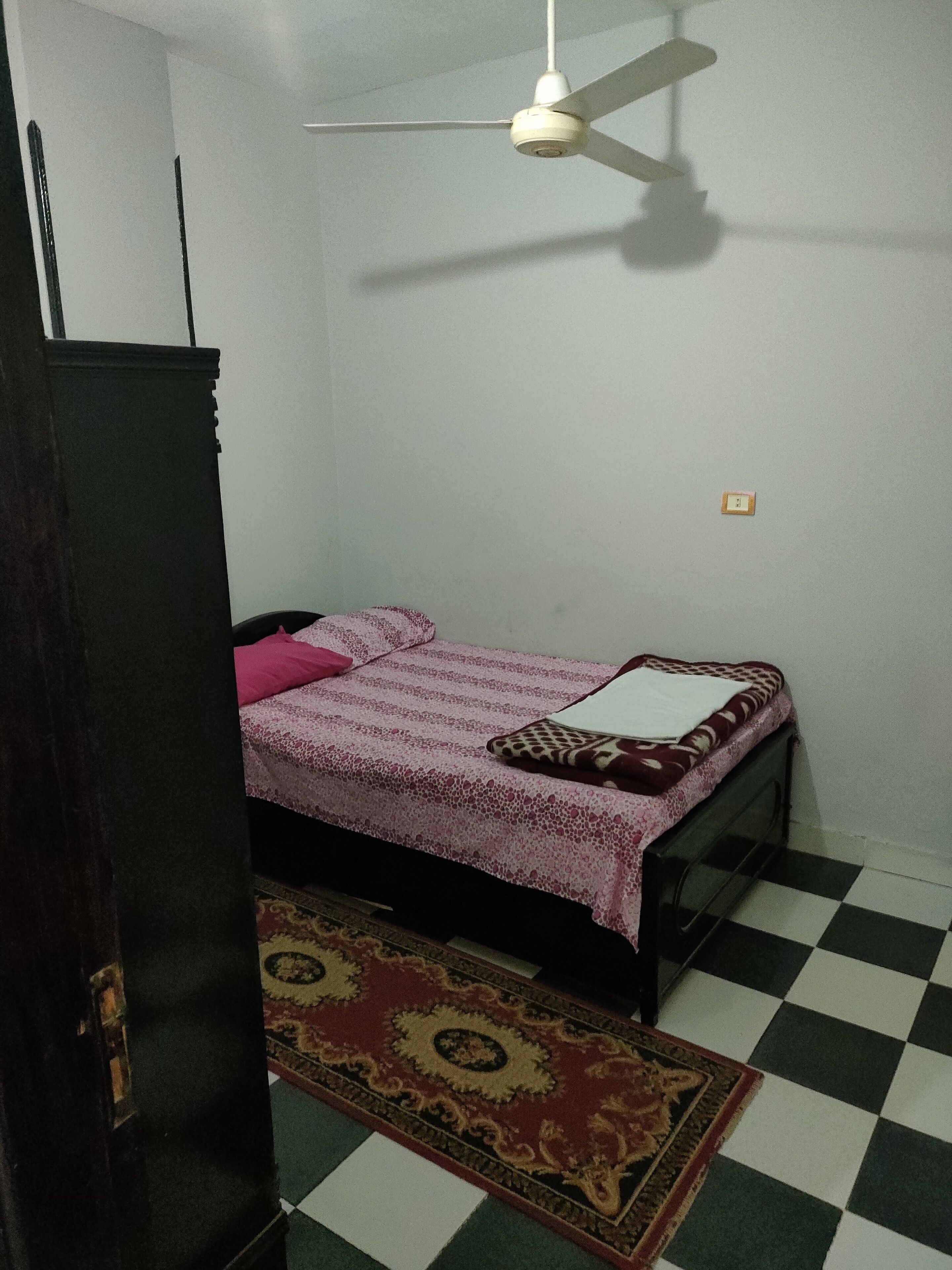 Een kleine, zwak verlichte kamer met een enkel bed, een plafondventilator en een kamerbreed tapijt met dessin.