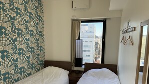 A hotel room with two beds, a window with a view of buildings, a wall-mounted air conditioner, and a coat rack. - HOTEL WAKO Tokyo Tree View (Tokyo)