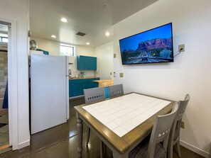 Cozinha compacta com armários cor turquesa, geladeira branca e área de refeições com mesa e cadeiras de madeira. Uma televisão de tela plana na parede exibe uma imagem panorâmica.
