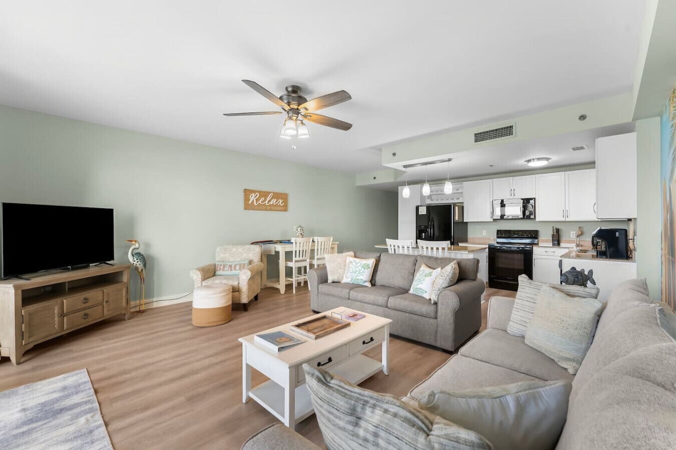 A modern living room with a flat-screen TV, a ceiling fan, a dining area with a table and chairs, and a kitchen with white cabinets.