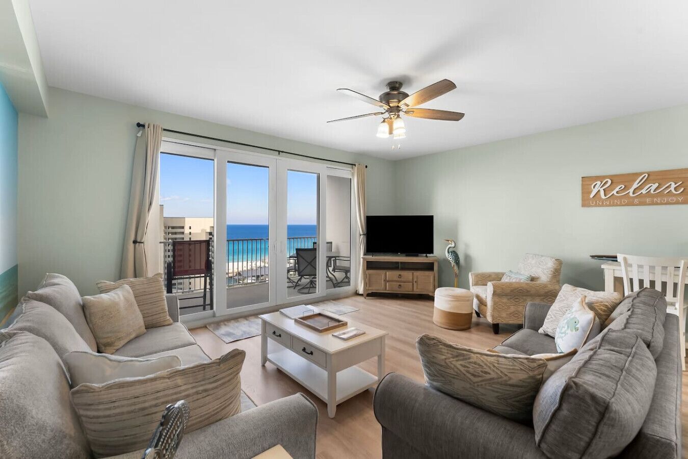 A spacious living room with a large sliding glass door leading to a balcony with a view of the ocean. There is a ceiling fan, a flat-screen TV, and a cozy seating area with a sofa and armchairs.