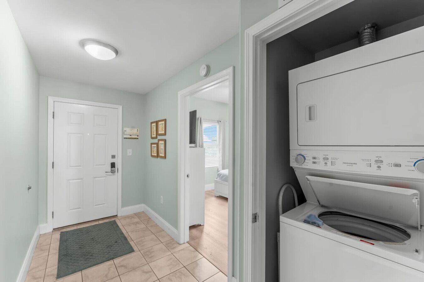 A laundry room with a front-loading washer and dryer, a door leading to a bedroom, and a small rug on the floor.