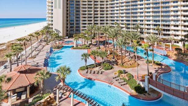 A resort with a large pool area, palm trees, and a beach in the background. - Hook Wine & Sinker - Beachfront Condo, 6th Floor (Panama City Beach)