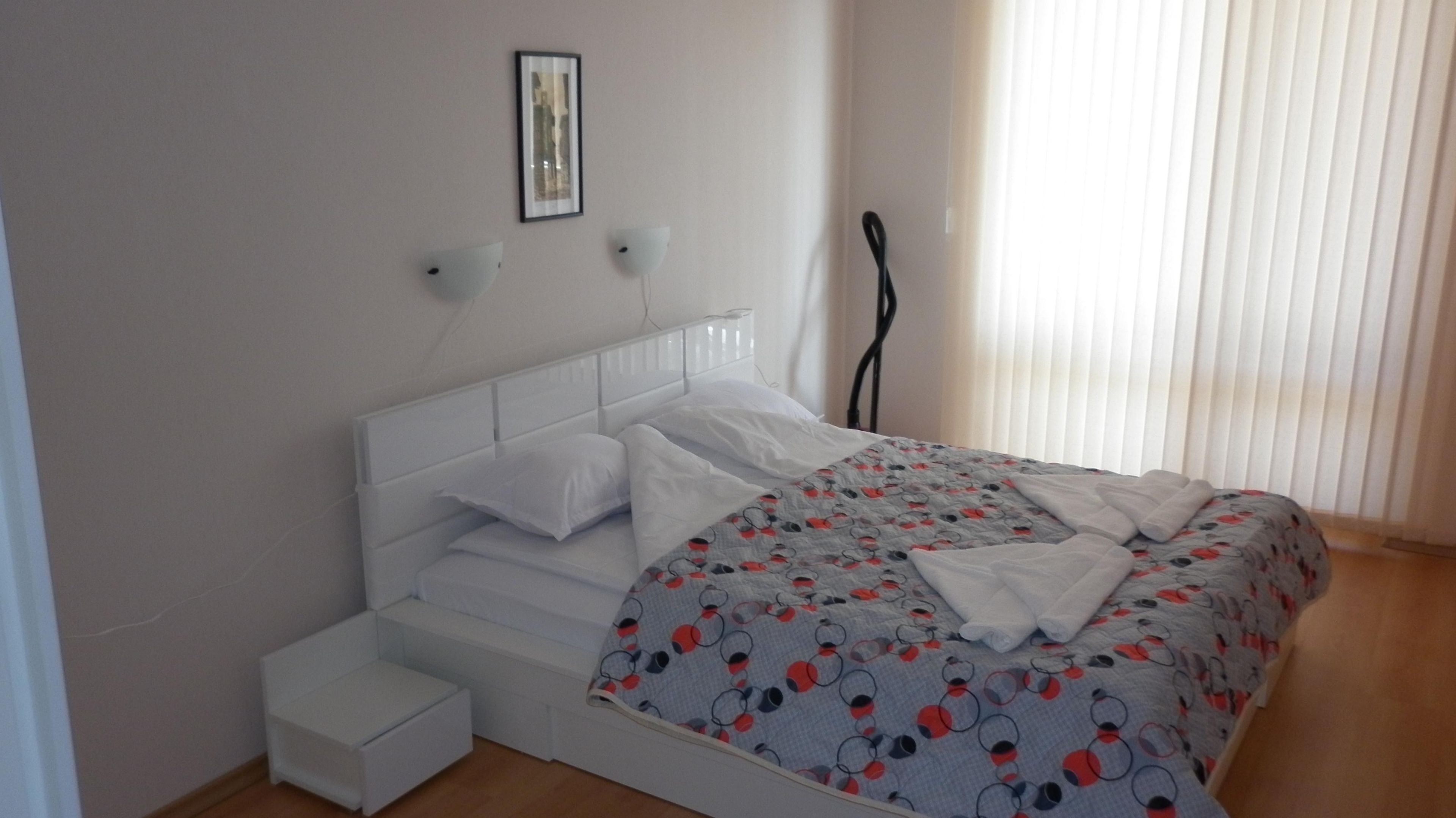 A bedroom with a white bed, a bedside table, and a window with blinds.