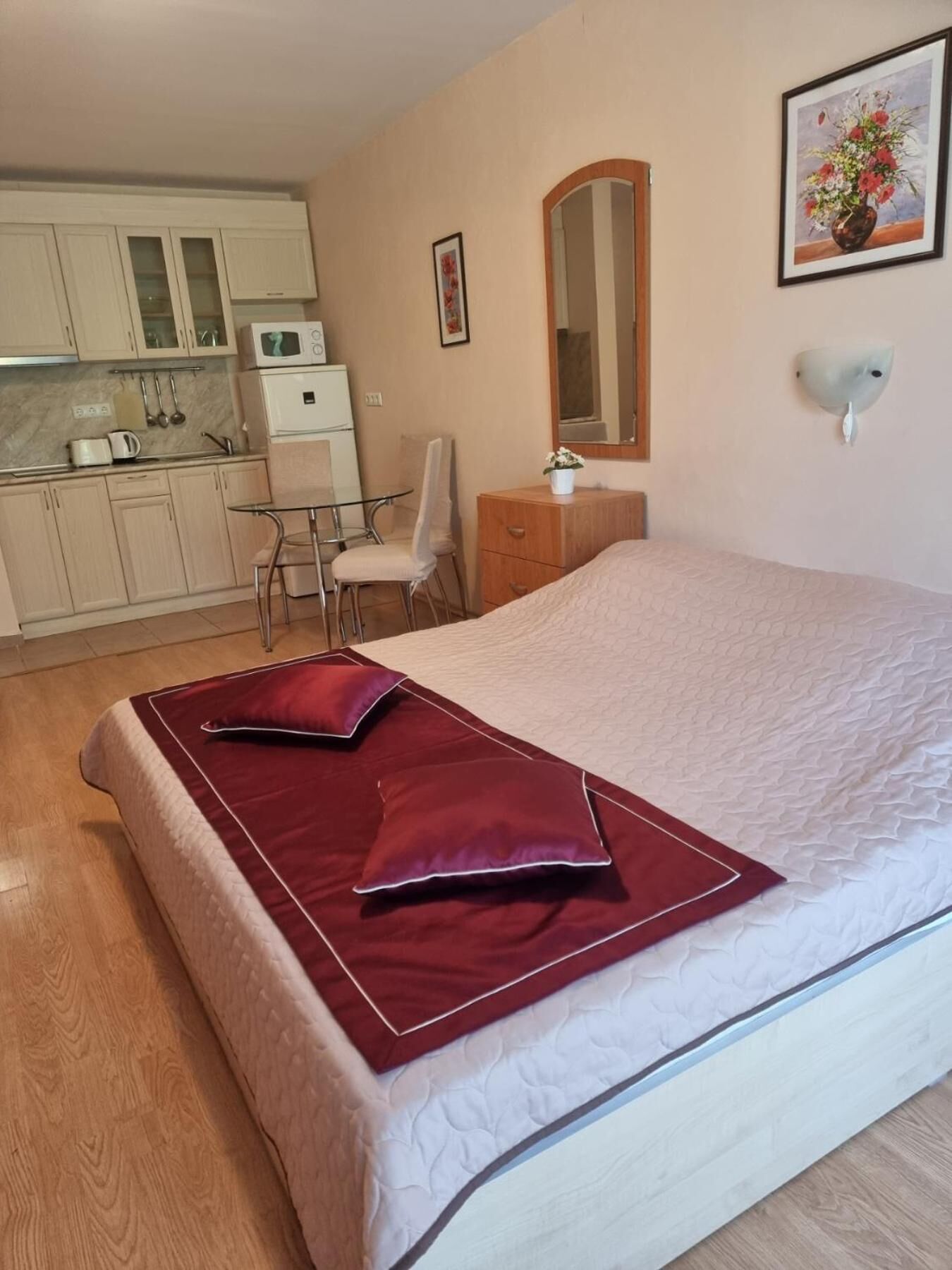A bedroom with a bed, a bedside table, a chair, a small desk, a microwave, and a kitchen area with cabinets and a sink.