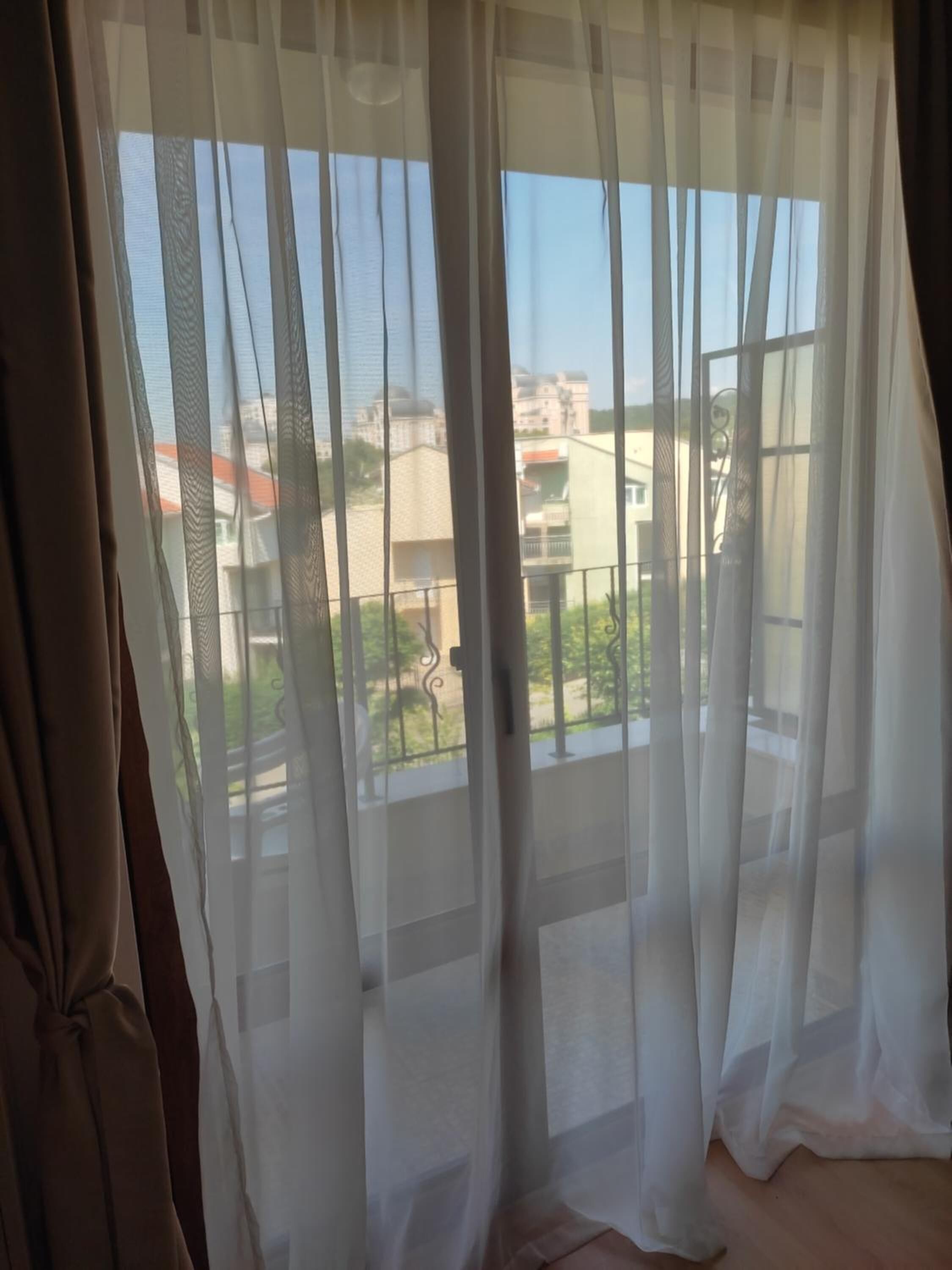 A room with sheer curtains, a balcony with a view of buildings and greenery.