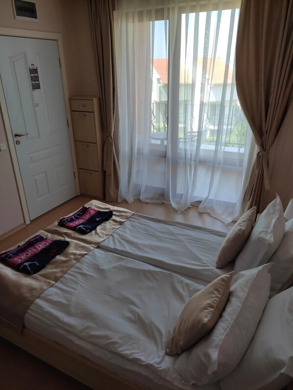 Une chambre avec deux lits, une porte et une fenêtre ornée de rideaux légers.