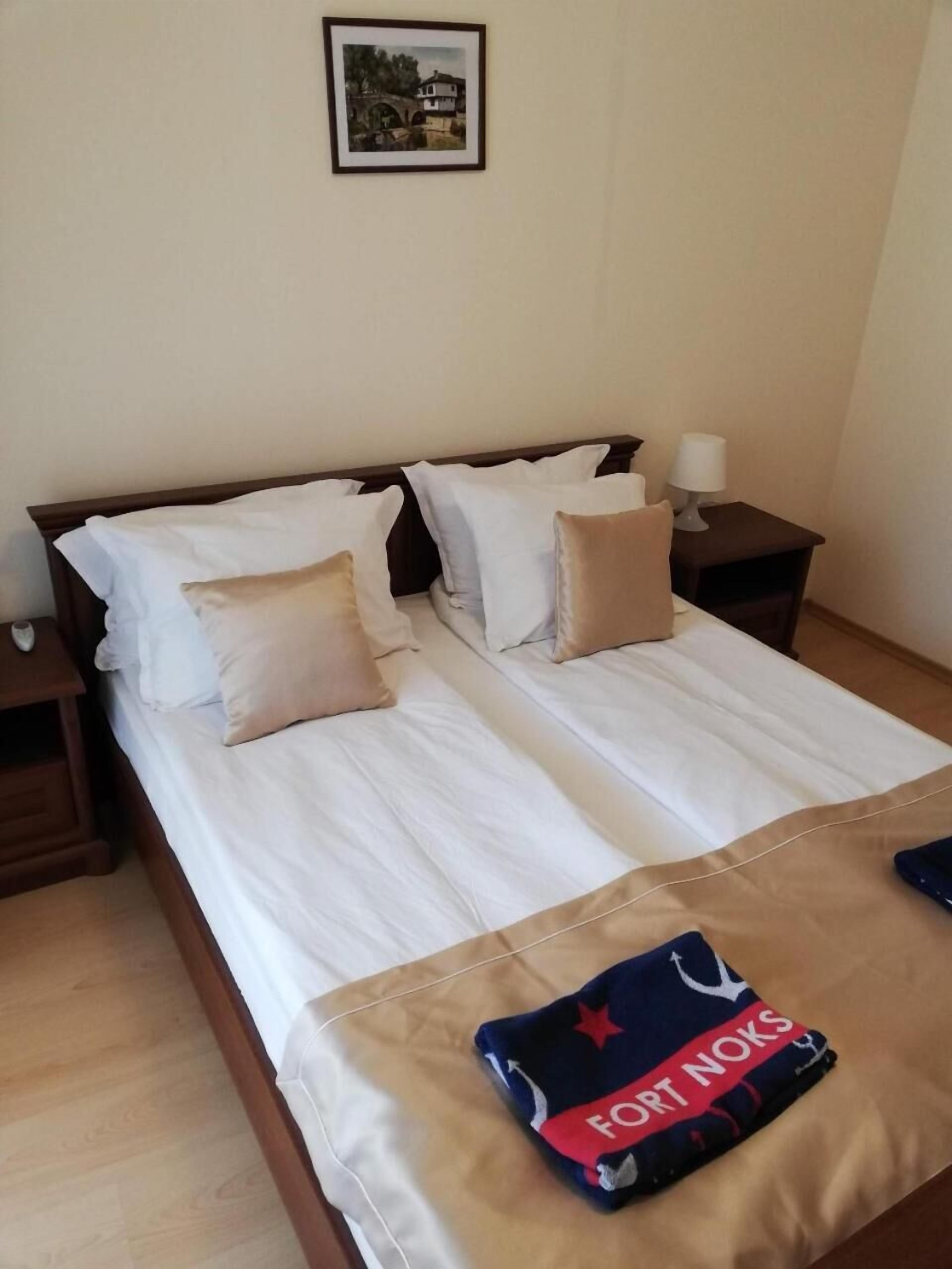A double bed with white linens, two pillows, a nightstand with a lamp, and a framed picture on the wall.