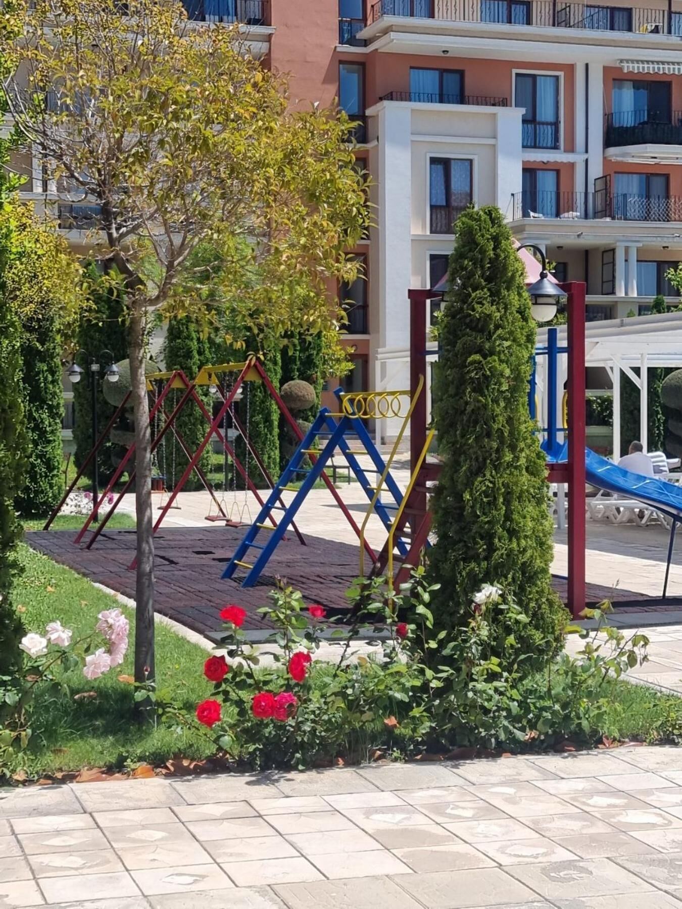 A playground with swings and climbing structures, surrounded by a garden with roses and a tall coniferous tree.