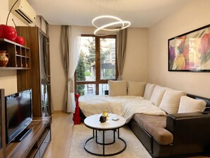 A modern living room with a sofa, a coffee table, a television, and a large window with curtains. - Prestige Fort Beach (Nessebar)