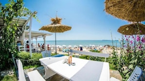 Una terraza con mesa y sillas, con vistas a una playa con sombrillas y gente.