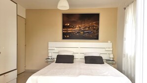 Un dormitorio moderno con una cama grande, un cabecero blanco, dos mesitas de noche con lámparas y un cuadro de paisaje urbano en la pared.