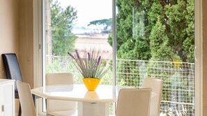 Un comedor moderno con una mesa y sillas blancas, un jarrón amarillo con flores moradas, y vistas a zonas verdes y un edificio a través de una puerta de vidrio.