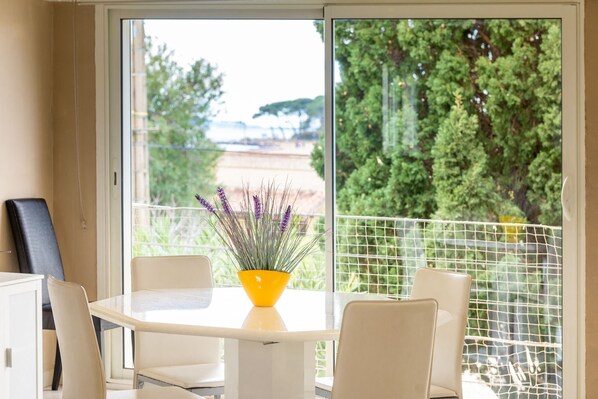 A modern dining area with a white table and chairs, a yellow vase with purple flowers, and a view of greenery and a building through a glass door.