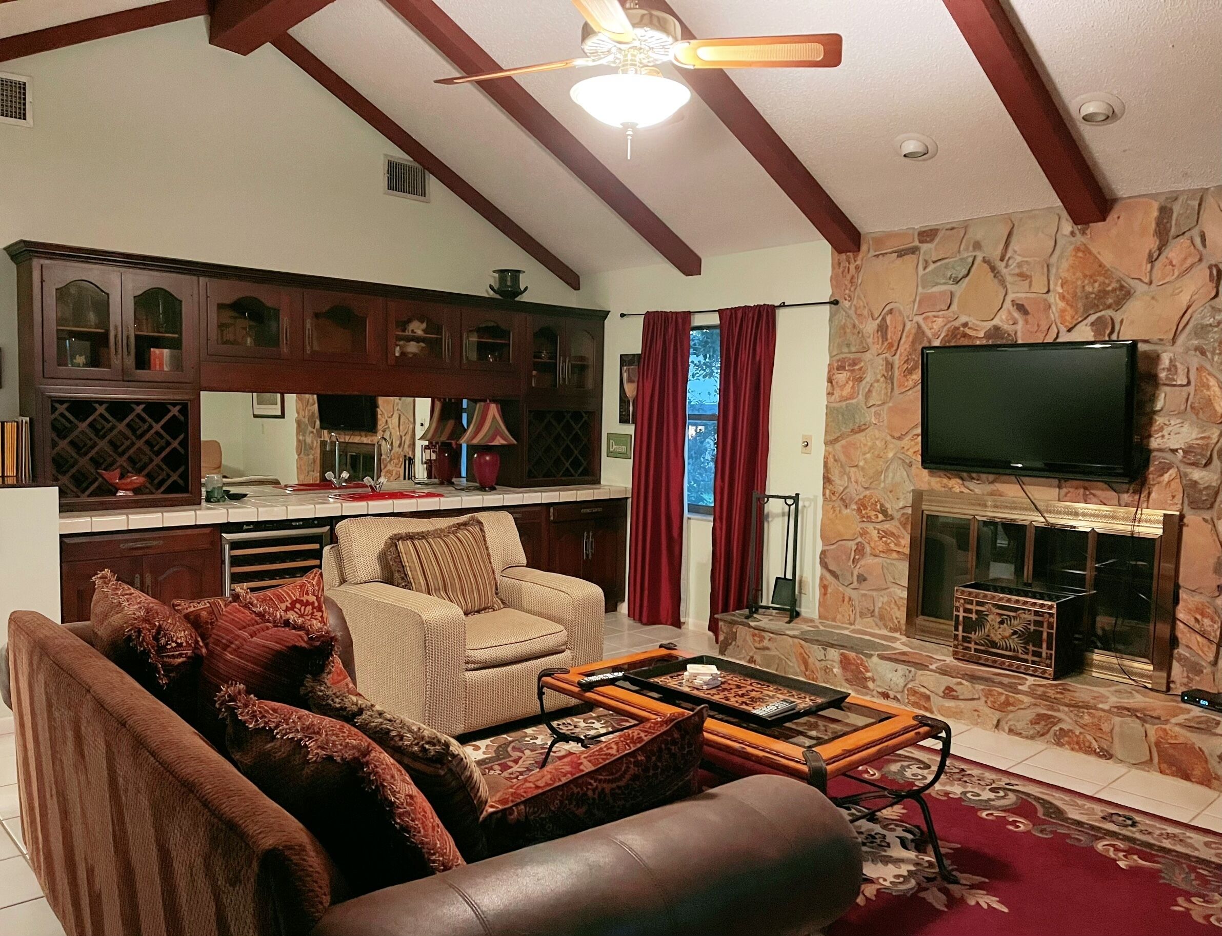 A living room with a sofa, armchair, and coffee table, a kitchen area with cabinets and a stove, and a stone fireplace with a TV mounted above it.