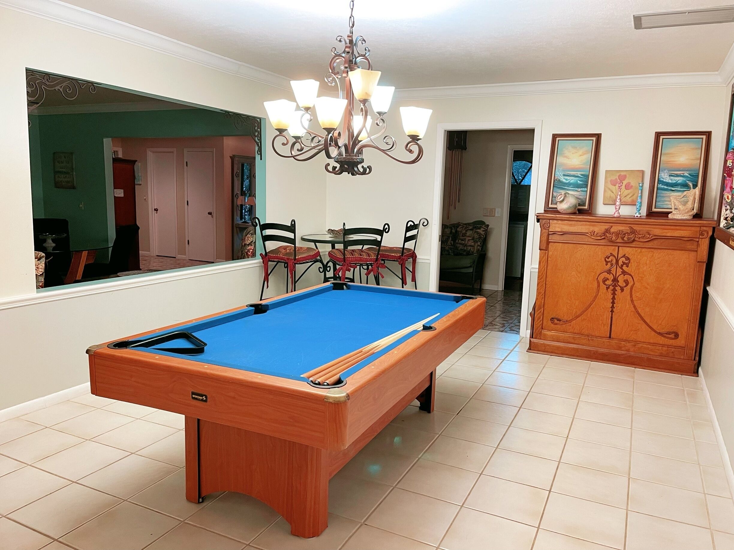 A pool table in a room with a chandelier, a wooden cabinet with artwork, and a dining area with chairs.