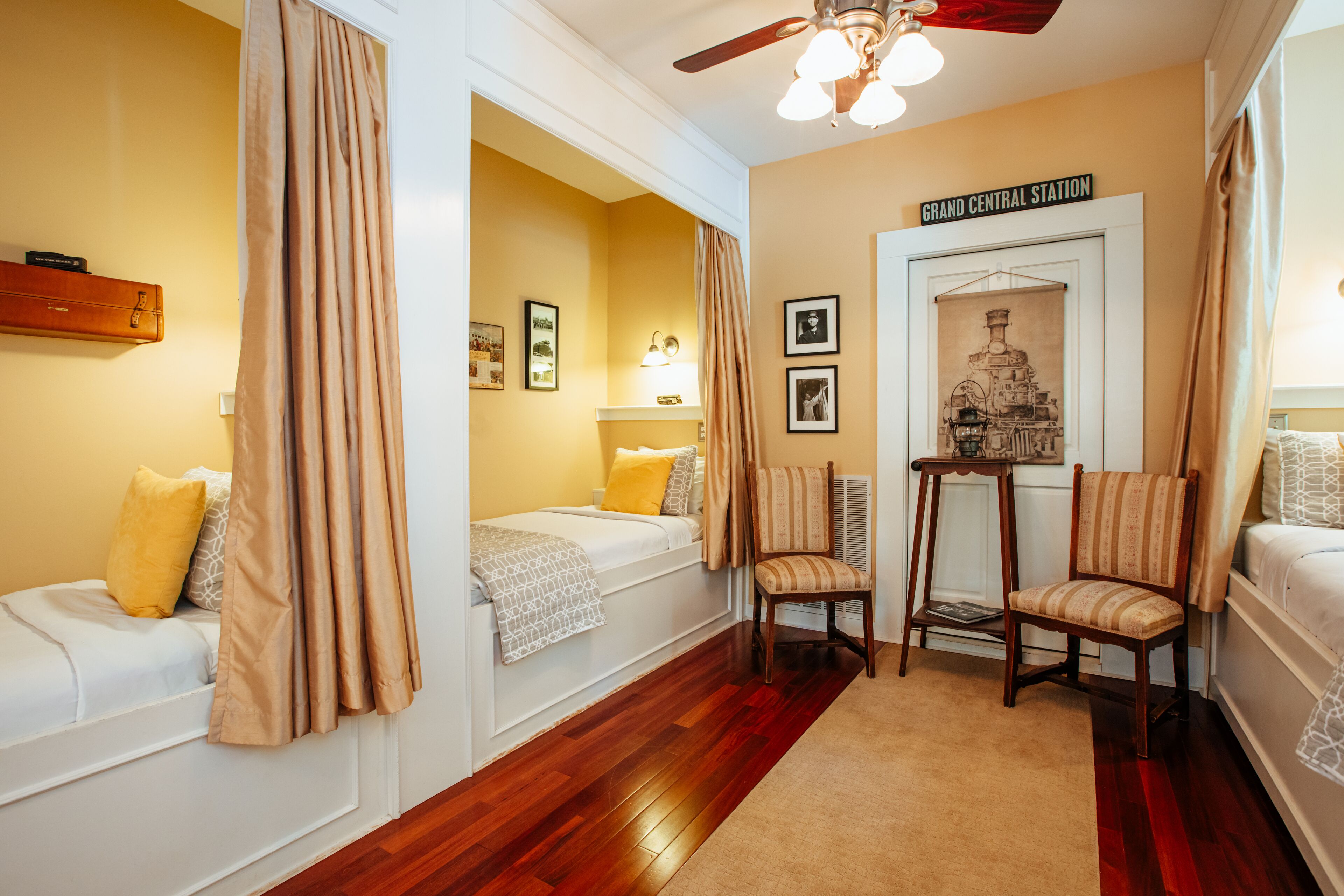 A hotel room with a bed, two chairs, a table, a ceiling fan, and a wall with a sign reading "Grand Central Station".