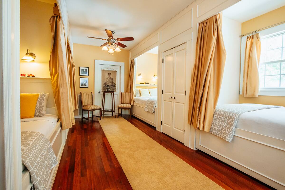 A hotel room with a wooden floor, two beds, a ceiling fan, and a small table with chairs.