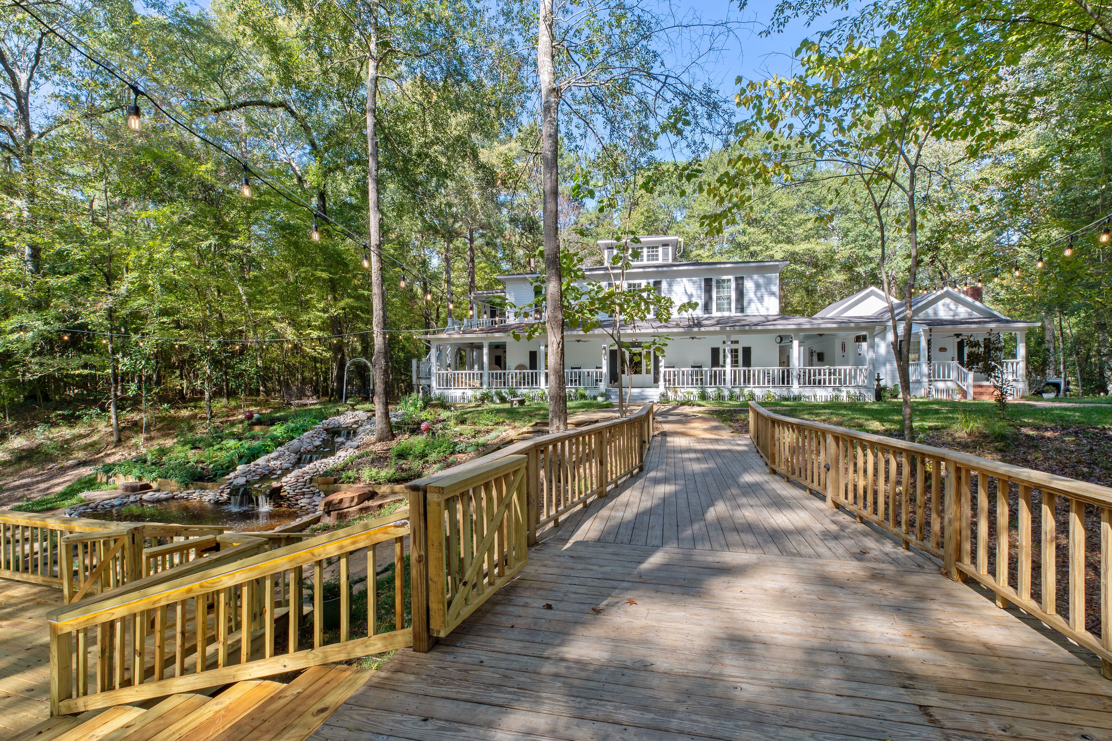 A wooden deck leading to a white house with a porch, surrounded by trees and a small pond.