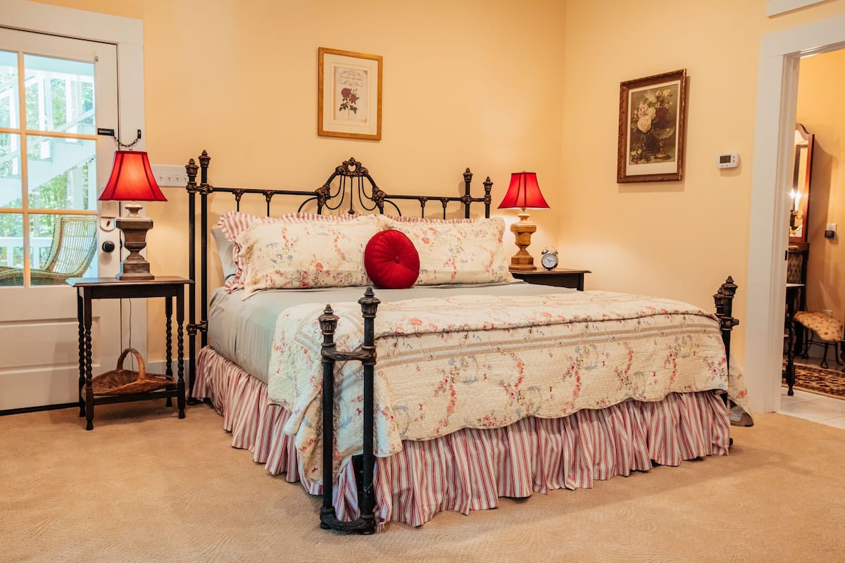 A bedroom with a four-poster bed, two bedside tables, a chair, and a door leading to another room.