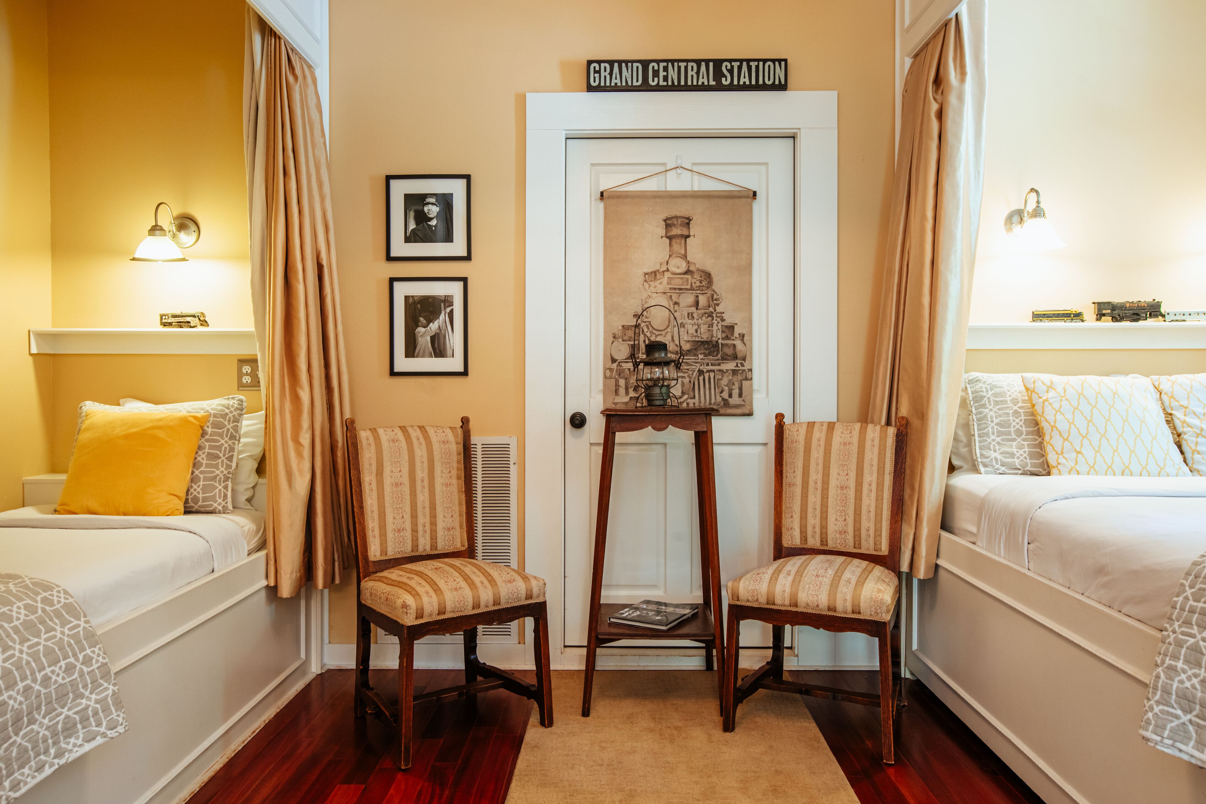 Habitación de hotel con dos camas, una silla de madera, una mesita y una puerta con el letrero 'Grand Central Station'.