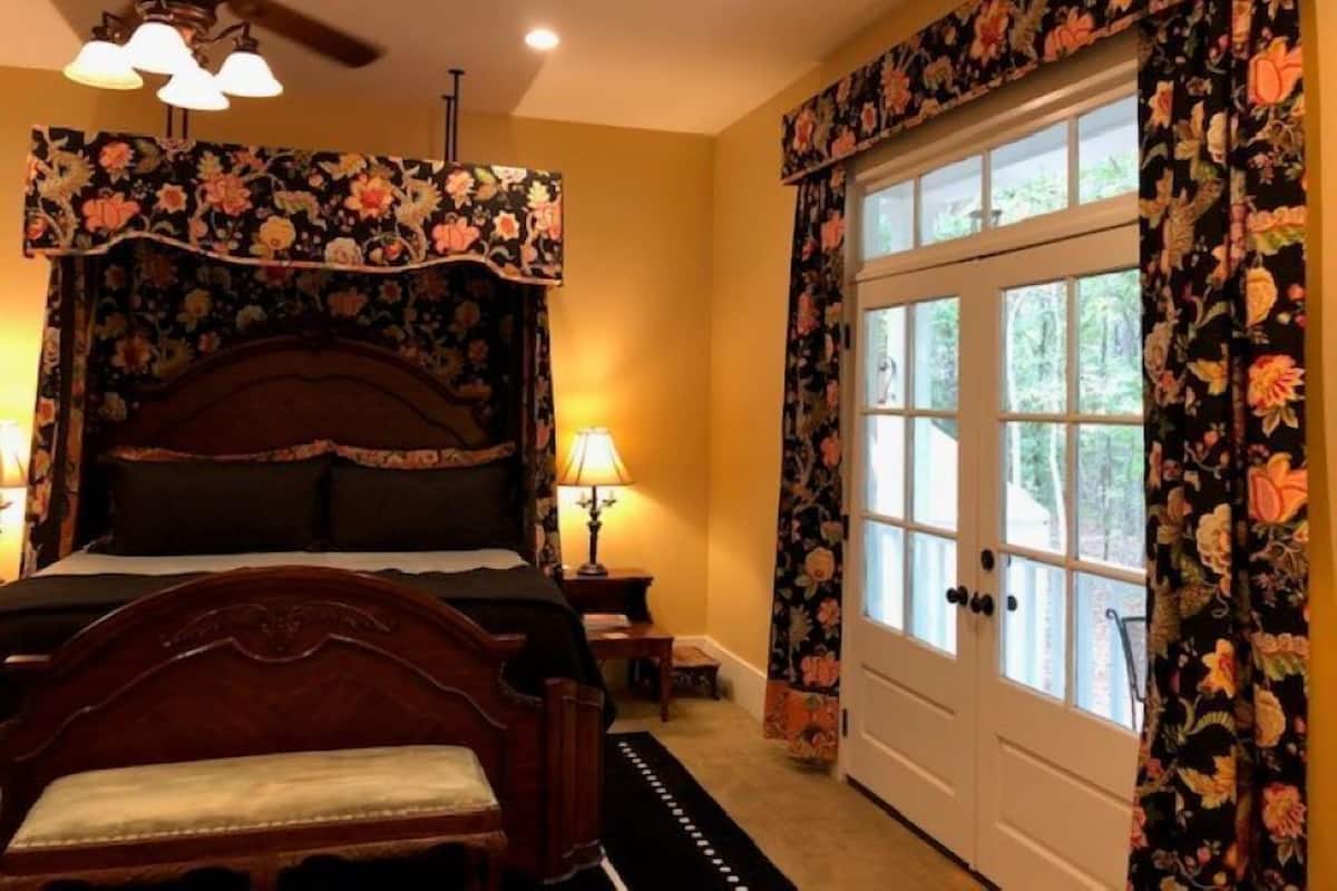 A bedroom with a four-poster bed, a ceiling fan, and a large window with floral curtains.