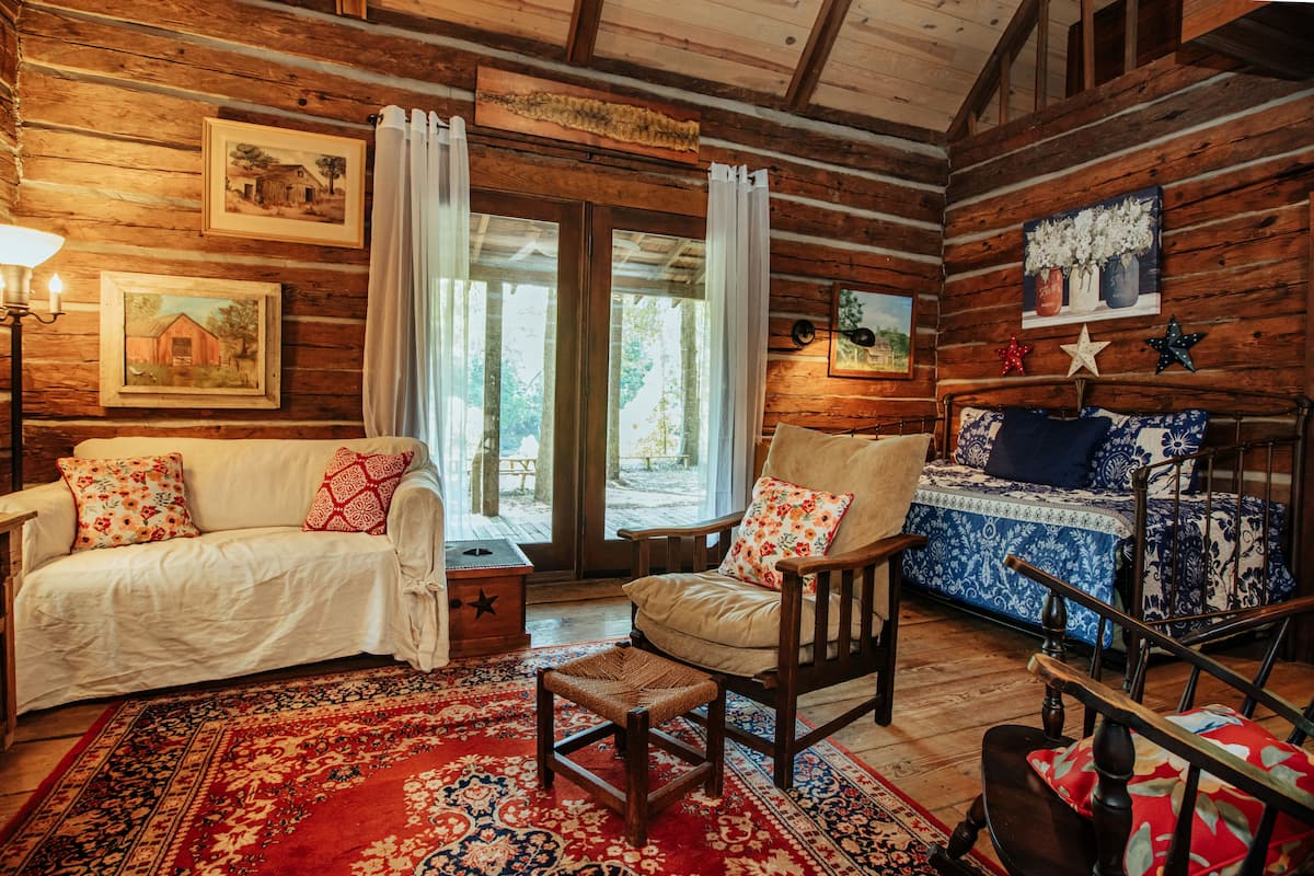 A log cabin interior with a wooden floor, a white sofa with patterned cushions, a rocking chair, and a bed with blue and white bedding.