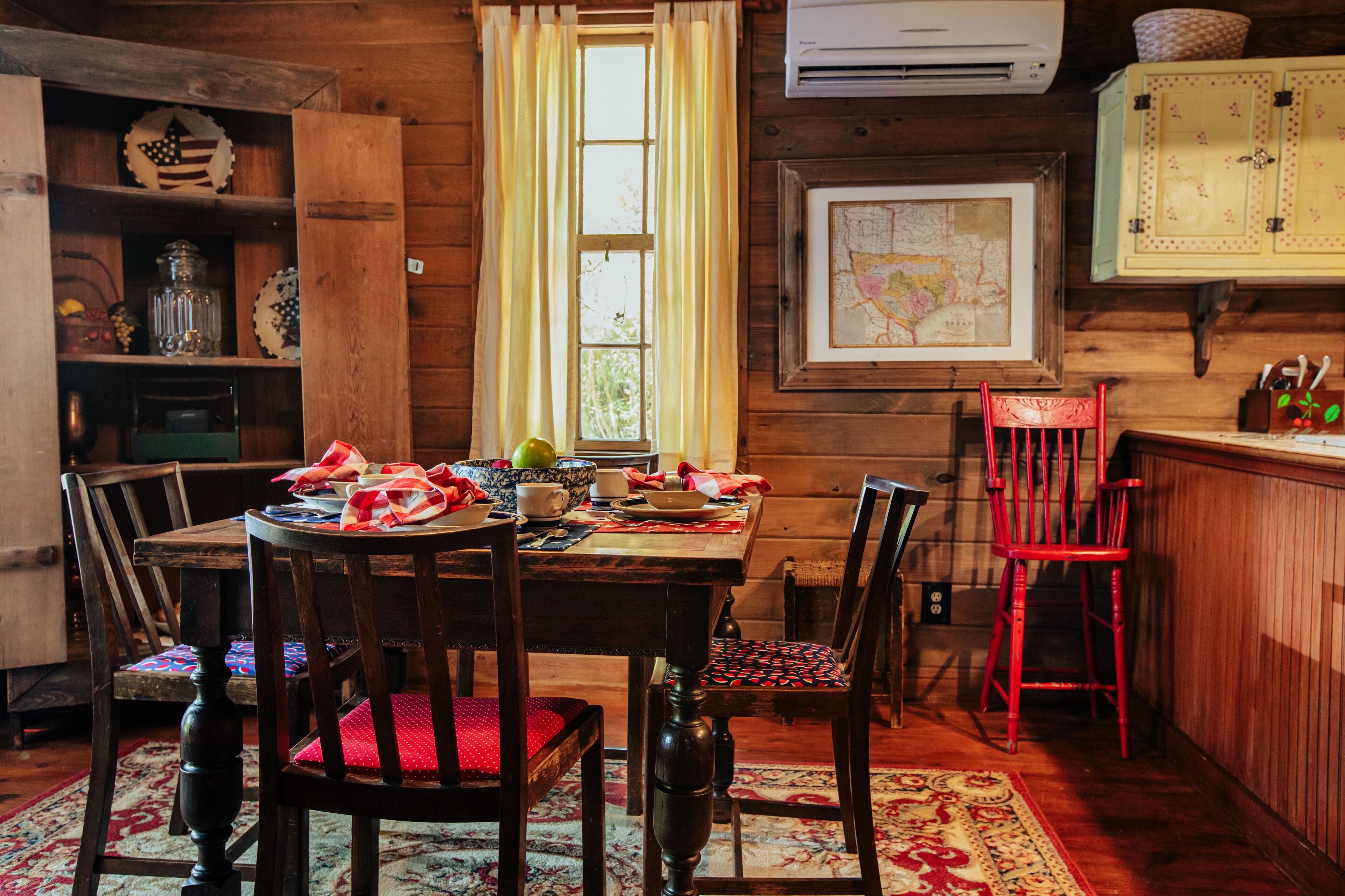 Interior de fusta amb una taula de dinar per a quatre, rodejada de cadires, un mapa a la paret i una finestra amb cortines.