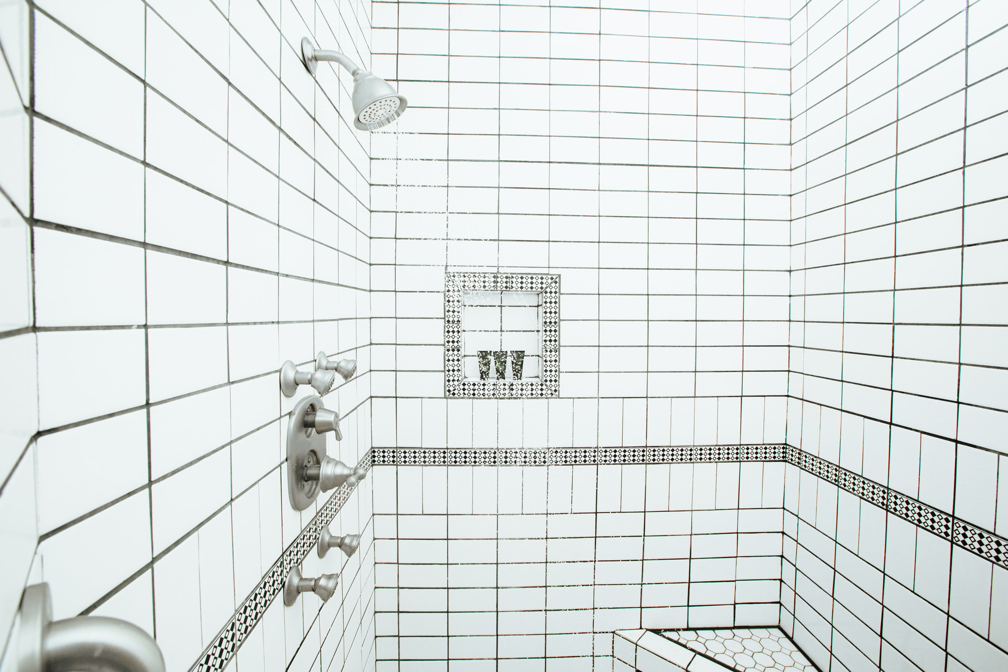 A bathroom with white tiled walls, a shower with a handheld showerhead, and a patterned tile border.