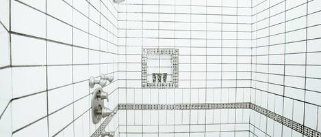 A bathroom with white tiled walls, a shower with a handheld showerhead, and a patterned tile border.