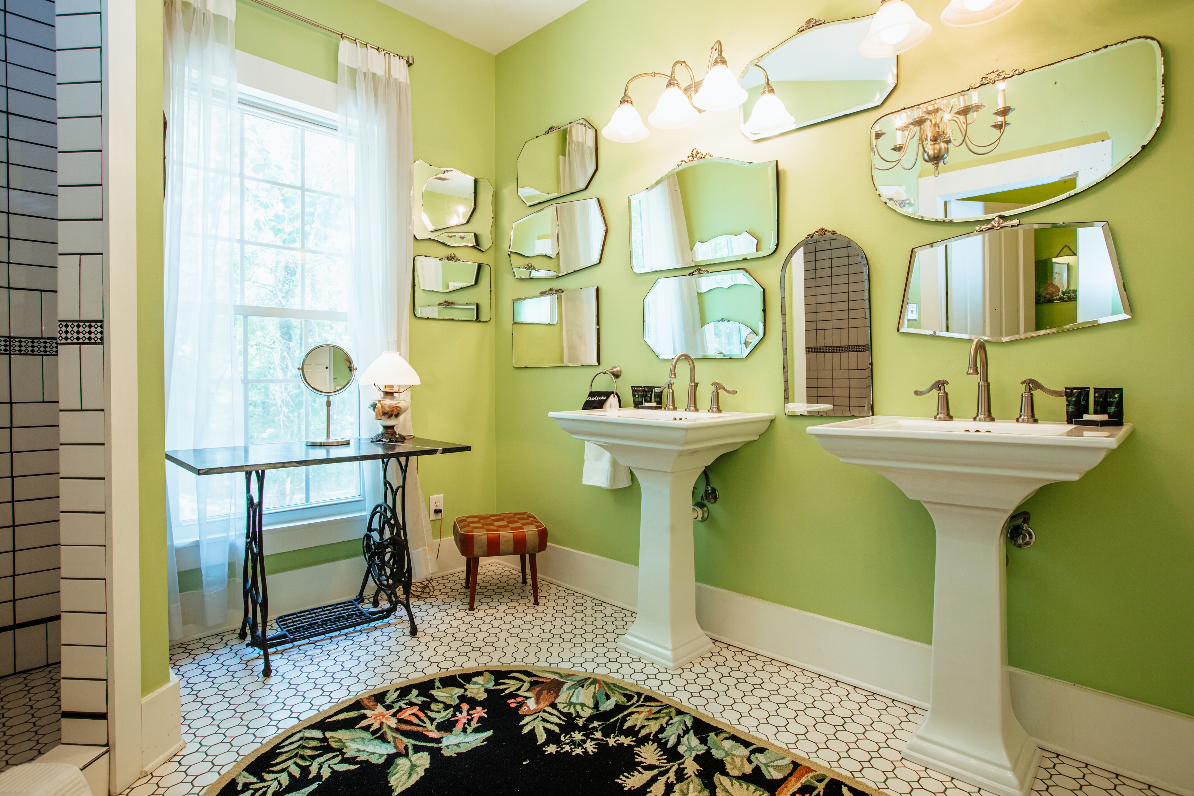 Een badkamer met groene wanden, twee wastafels, meerdere spiegels, een naaimachine en een vloerkleed met dessin.