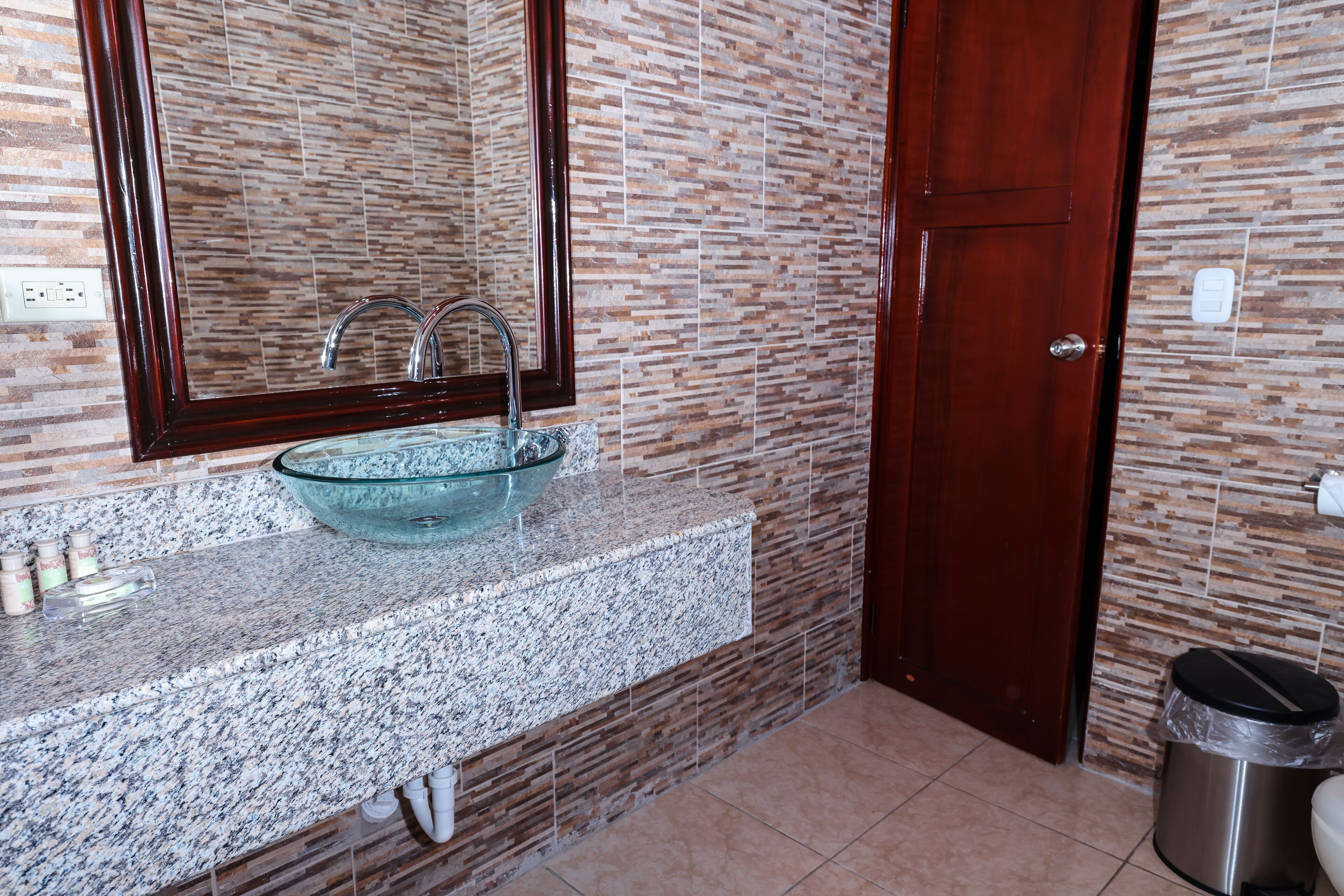 A bathroom with a granite countertop, a glass bowl sink, a large mirror, and a wooden door.