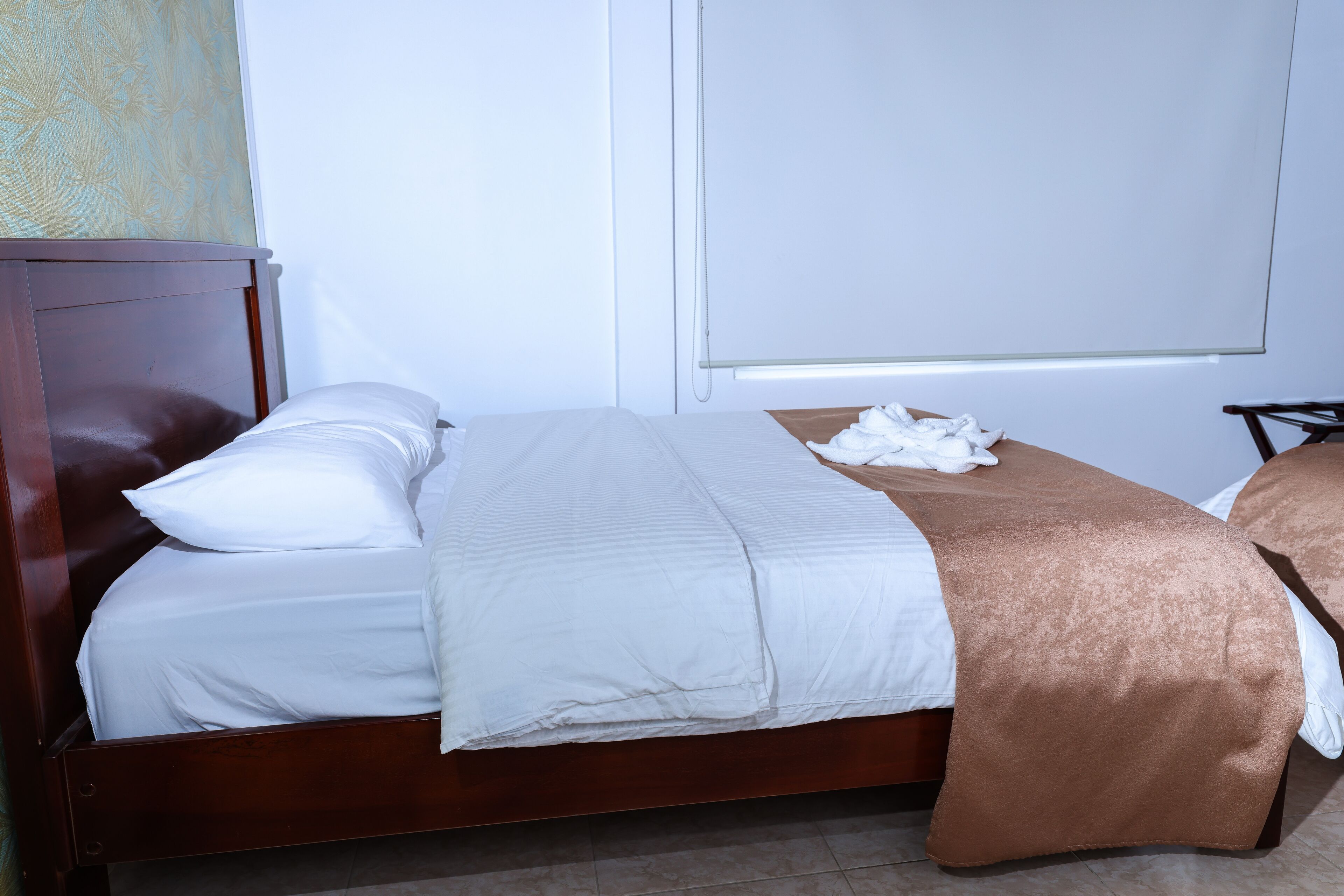 Una cama bien tendida con sábanas blancas y un plaid marrón, un cabecero de madera y una mesita de noche con una lámpara blanca.
