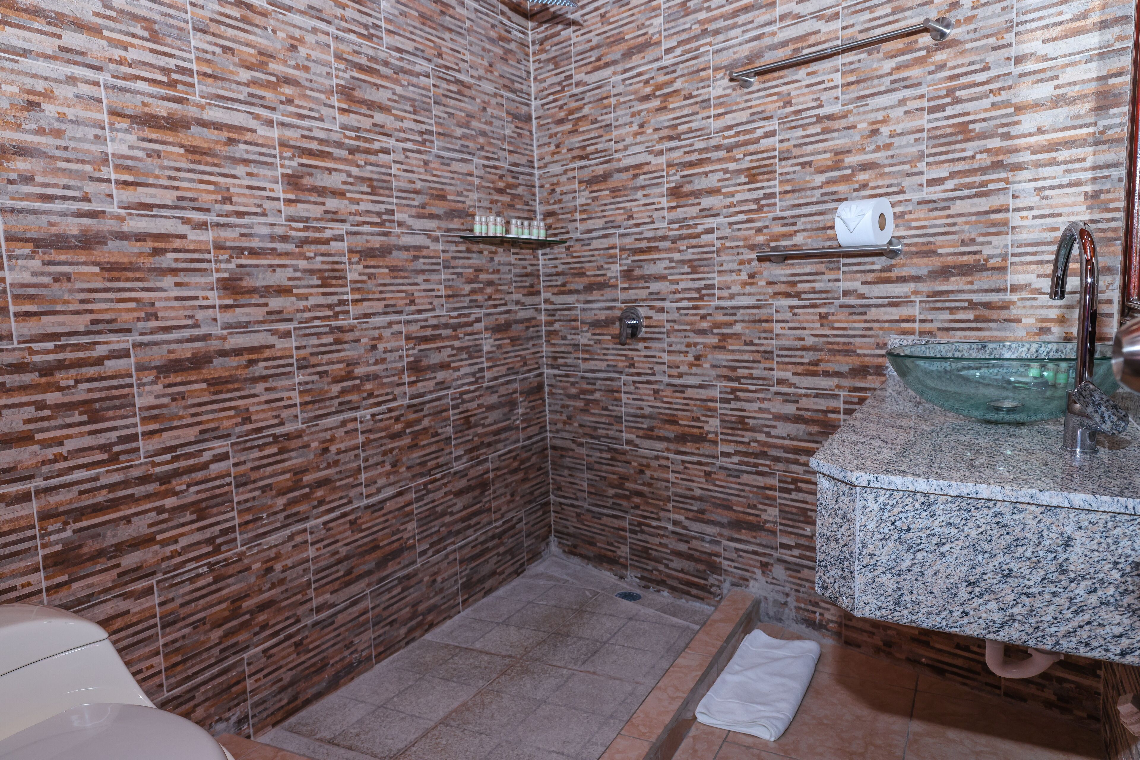 Un baño con zona de ducha, encimera de granito y lavamanos de vidrio.