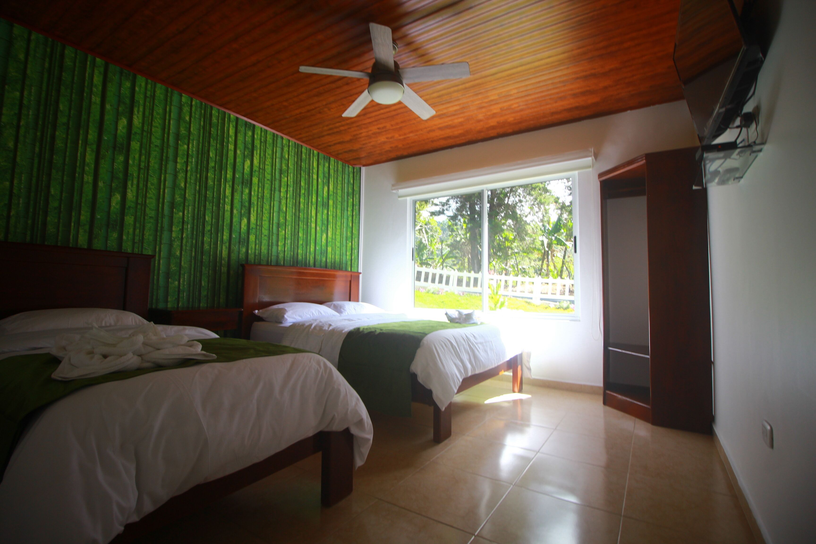 Quarto de hotel com duas camas, ventoinha no teto, grande janela com vista para áreas verdes e teto de madeira.
