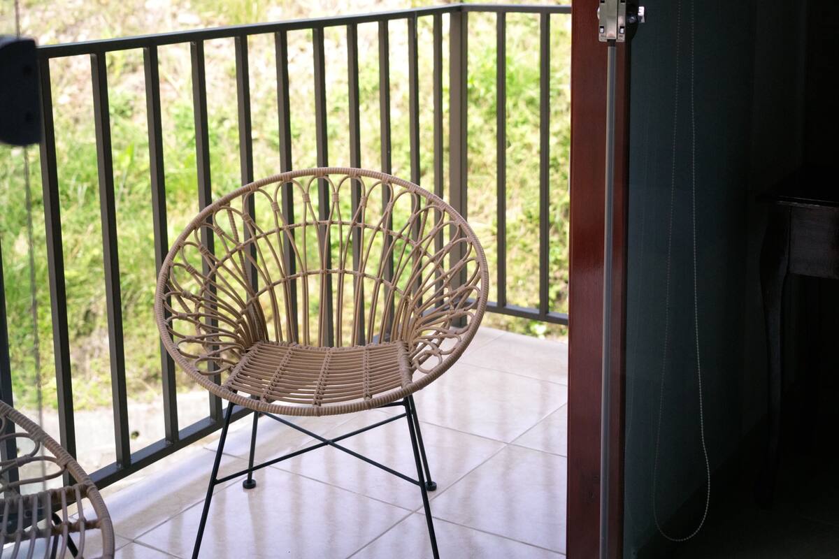 Un balcón con una silla de mimbre y una mesa, con vistas a un jardín con árboles y césped.