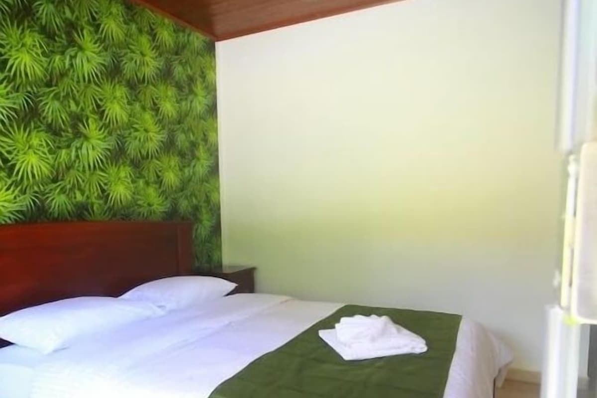 Una cama individual con colcha verde y sábanas blancas, cabecera de madera, ventilador de techo y pared con estampado de hojas verdes.