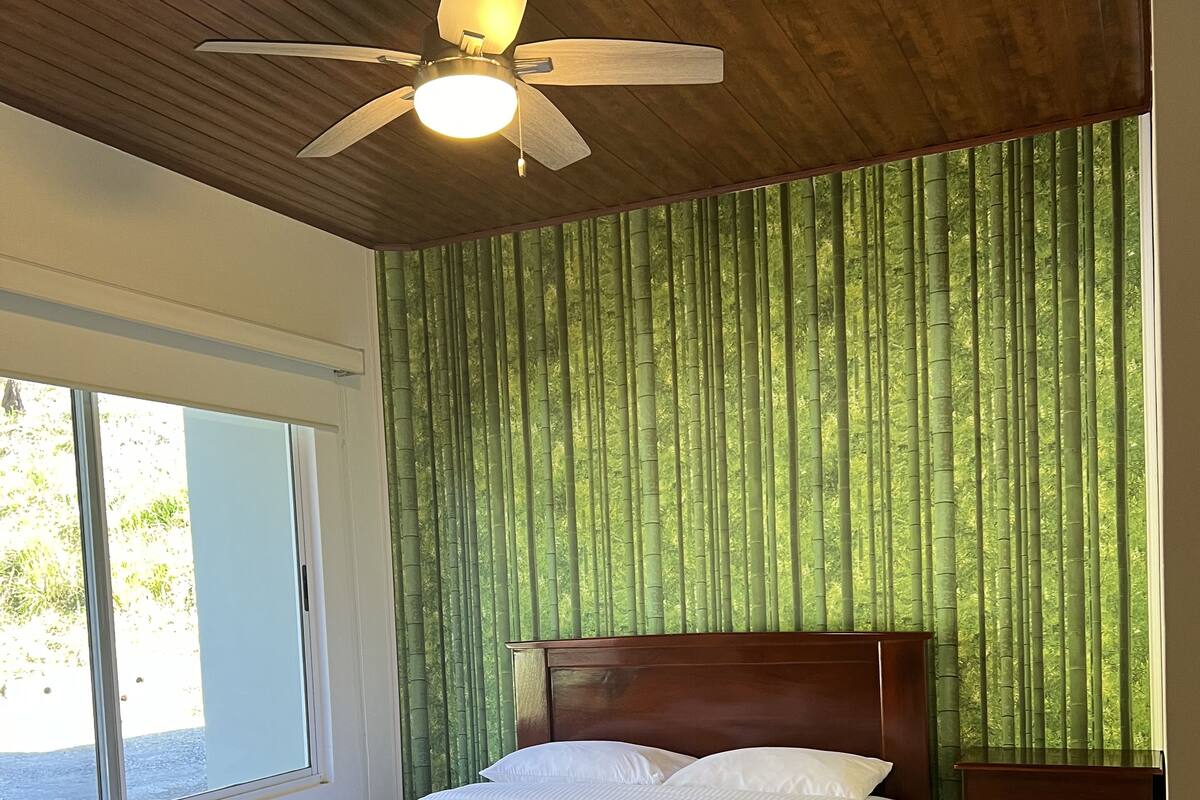 Un dormitorio con cama, ventilador de techo, ventana y vista a bambú desde afuera.