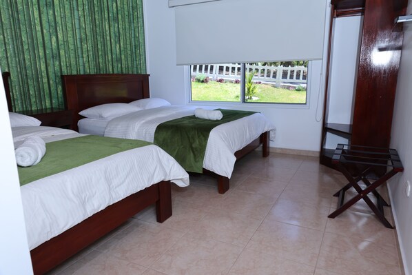 A hotel room with two beds, a window with a view of a white fence and greenery, and a wooden chair.