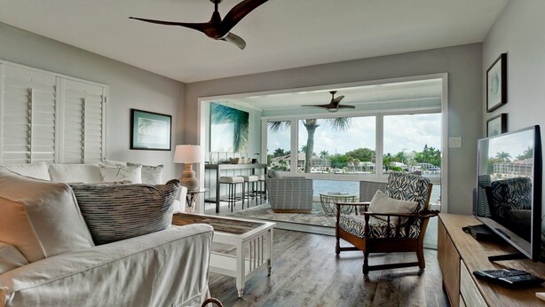 A spacious living room with a large window, a ceiling fan, a TV, and a dining area with a view of the water.