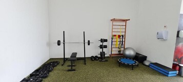 Uma pequena academia com barra de exercícios, banco, halteres, kettlebell, escada e um trampolim.