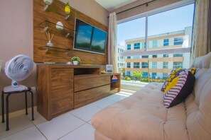 A modern living room with a sofa, a TV mounted on a wooden wall, a fan, and a view of a building through a window.