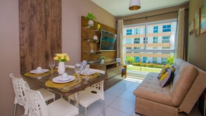 A dining area with a wooden table set for four, white chairs, a sofa, and a TV mounted on a wooden panel. - Flat em Porto de Galinhas - Muro Alto Clube (Ipojuca)