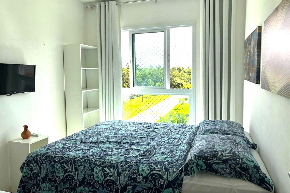 Une chambre à coucher avec un lit, une télévision, une fenêtre avec des rideaux et une vue sur la verdure.
