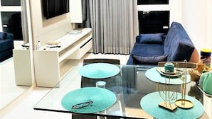 A modern living room with a glass dining table, two blue chairs, a sofa, a TV mounted on the wall, and a grey curtain. - Flat em Porto de Galinhas - Muro Alto Clube (Ipojuca)