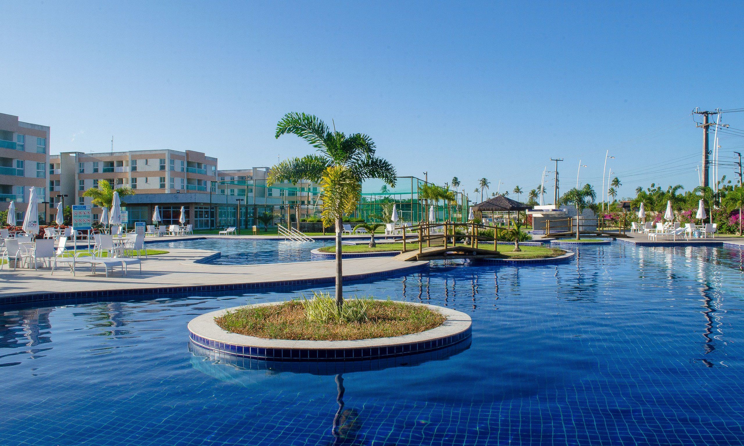 Una piscina con sillas de descanso y sombrillas, rodeada de palmeras y un edificio al fondo.